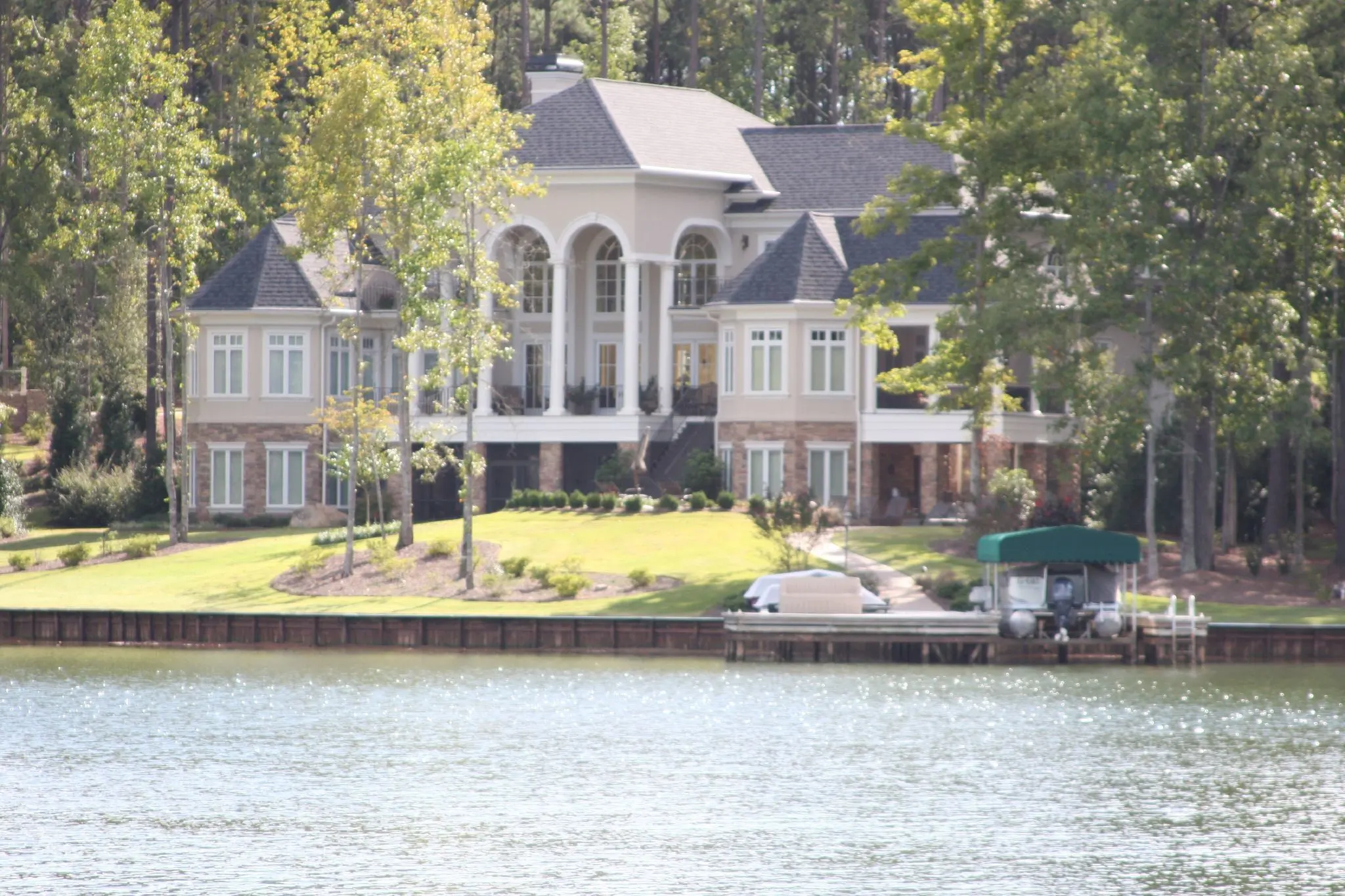 A large lakefront home among hardwoods and pines with a covered boat dock on a seawall at Lake Oconee.