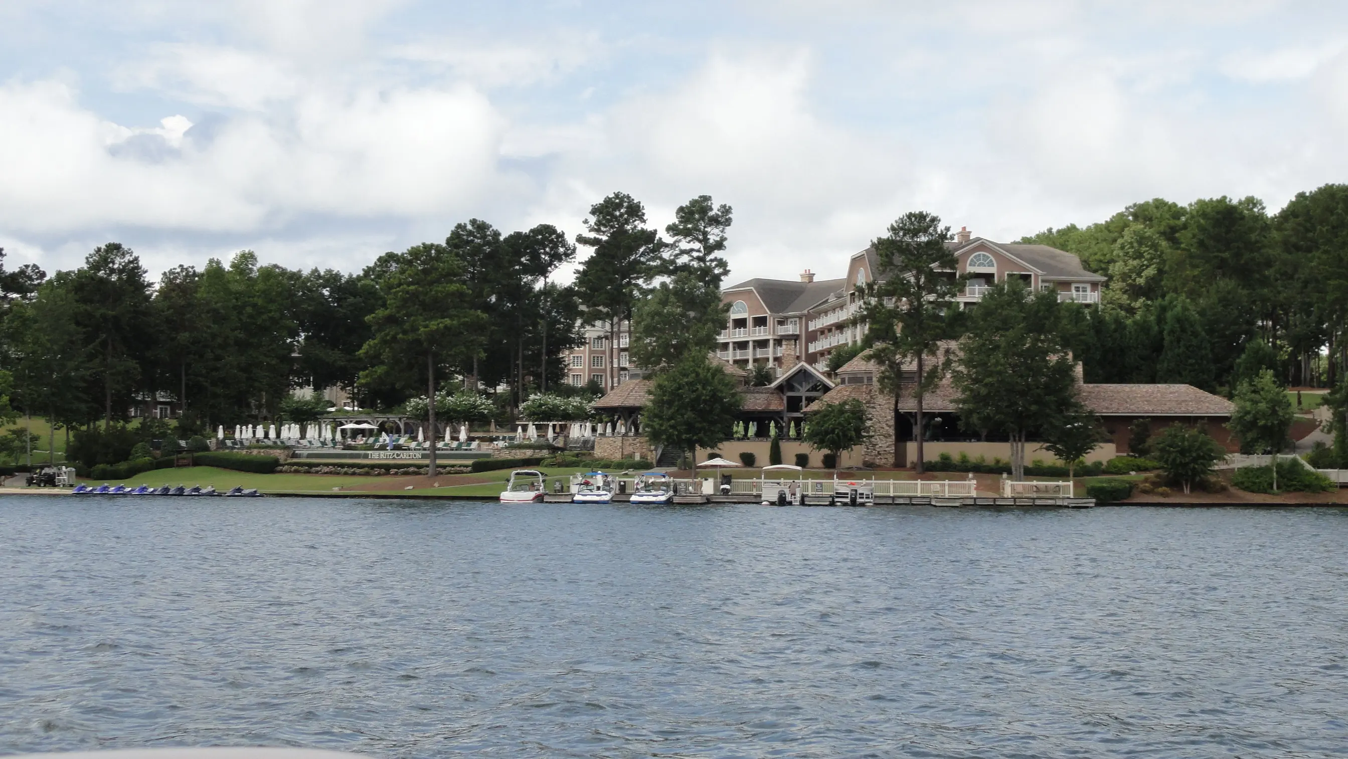A pine-lined Lake Oconee shoreline with a lakeside lodge, lawn and docks under a soft cloudy sky.