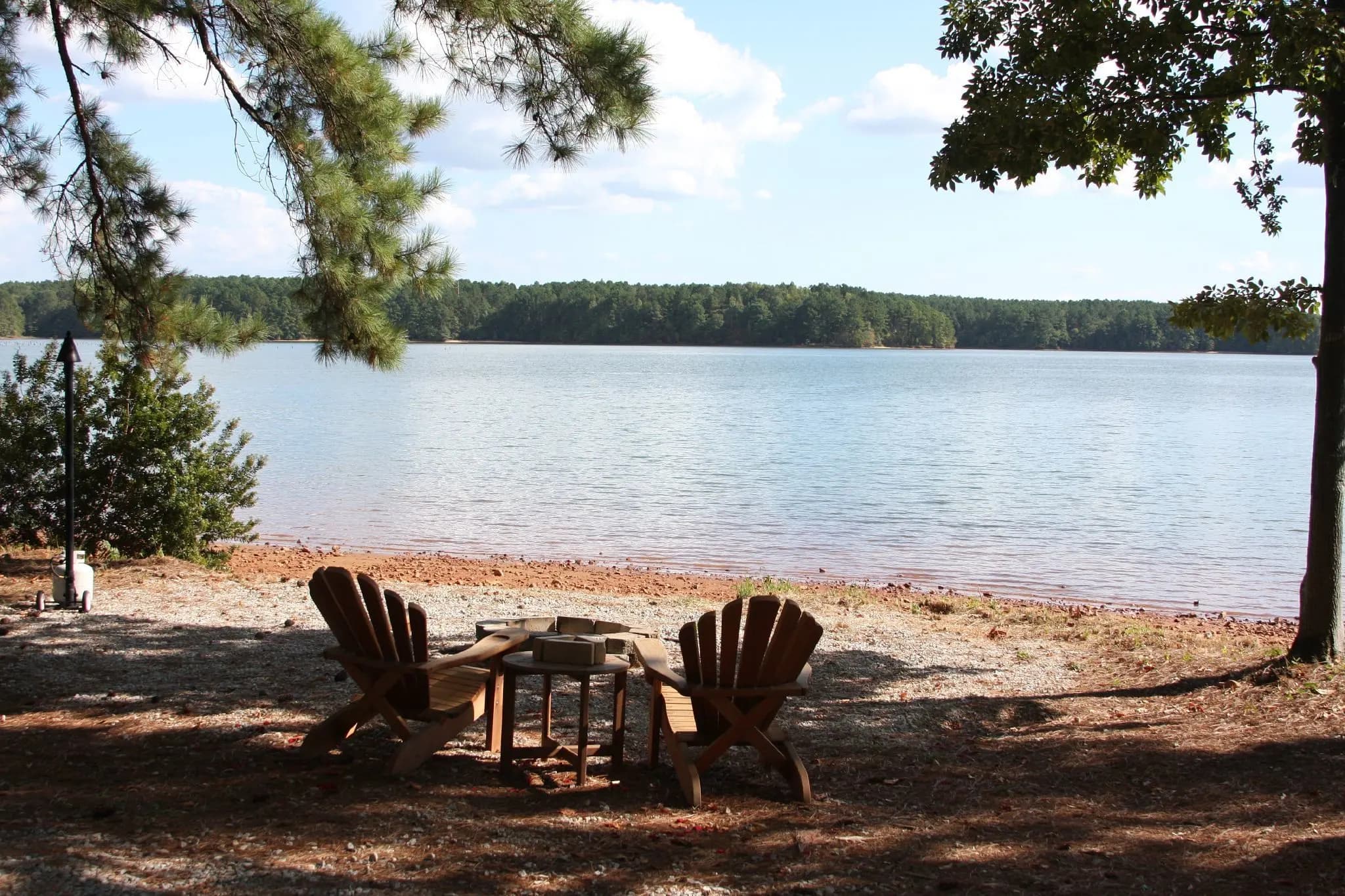 Two Adirondack chairs on a pine-shaded sandy shore looking out over Lake Oconee.